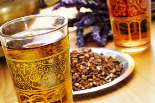 close up of glass of herbal tea and dried herbs on a plate nearby and purple flowers nearby