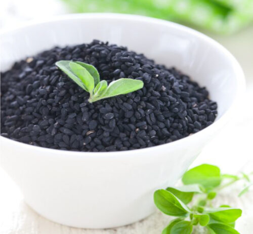 a white bowl filled with black seeds and some green leaves lie on top and a few leave scattered around bowl