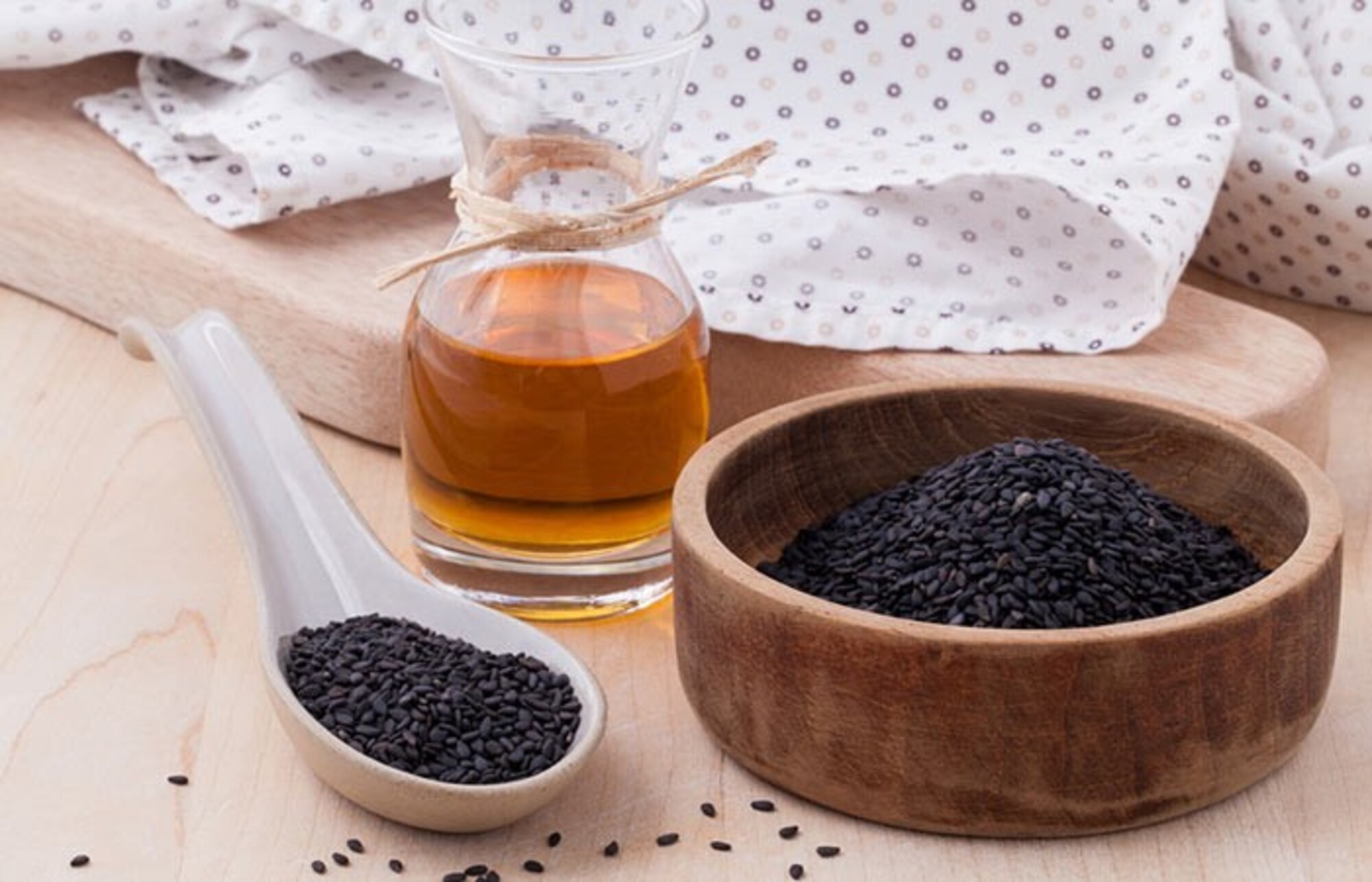 a brown wooden bowl and white spoon filled with black seeds and a bottle of black seed oil nearby- both on a wooden surface with a spotted piece of material in background