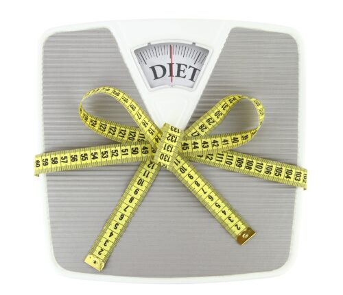 close up of scales with the words DIET in the window and yellow measuring tape in a bow shape around it