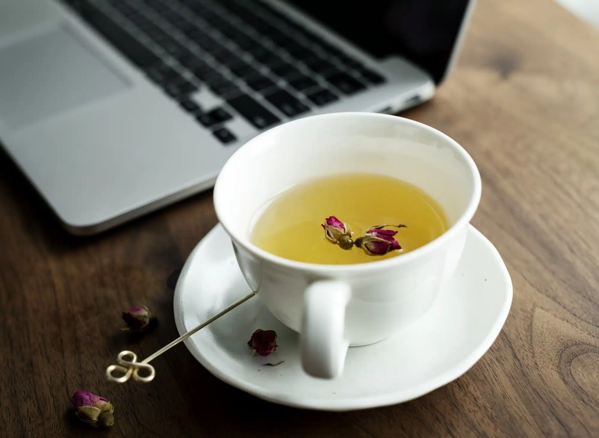 white cup with herbal tea on saucer with red dried flower floating on top, nest to an open laptop on a wooden desk