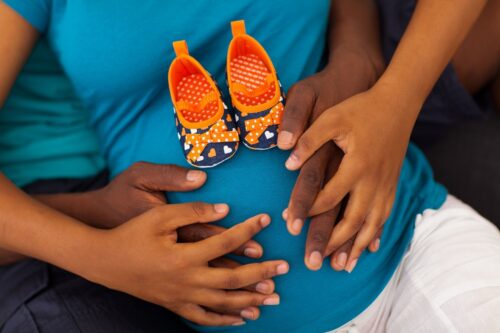 close up of couples hands embracing her pregnant belly with a pair of orange and blue baby shoes on her blue top