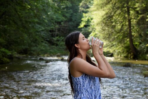 Woman drinking a glass of water and standing near a river in nature