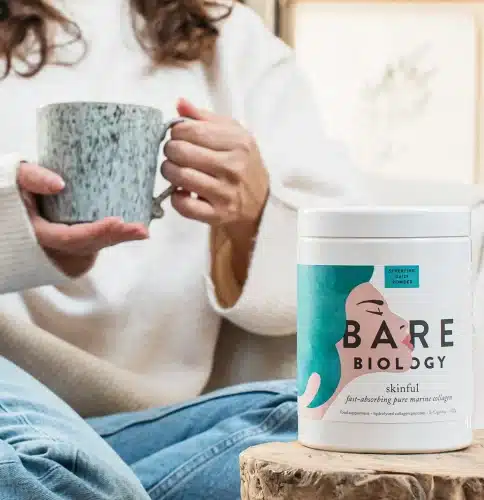 woman in background holding a cup in both hands and tub of Bare Biology powder in foreground