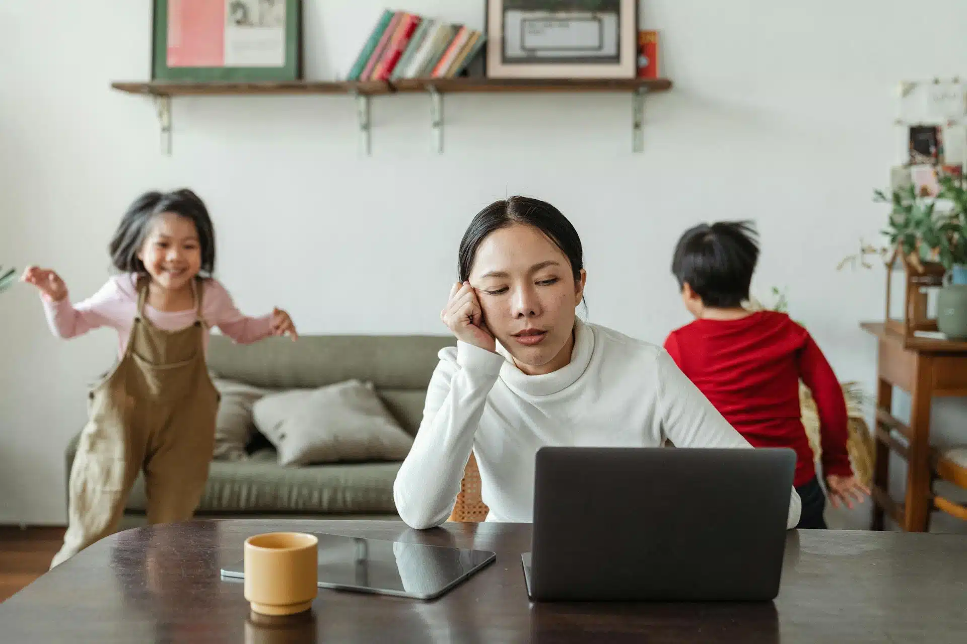 A tired trying to do work on her laptop while her two kids are playing in the background